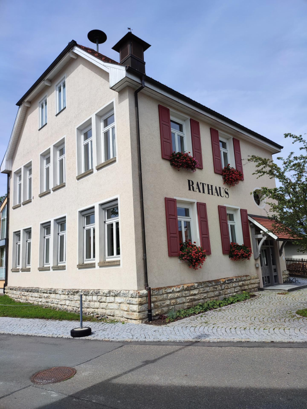 Das Rathaus-Hotel, ein weißes Gebäude mit roten Fensterläden, umgeben von Bäumen, Pflanzen, Blumen und Gras, mit einer Straße unten und dem Himmel im Hintergrund.