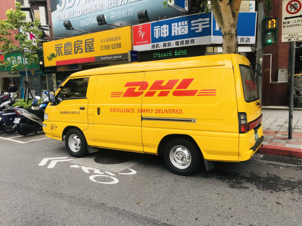 Ein gelber DHL-Lieferwagen steht auf einer Straße neben Gebäuden, Bäumen, Schildern, Pfählen, Ampeln und einem Fußgänger auf dem Bürgersteig.
