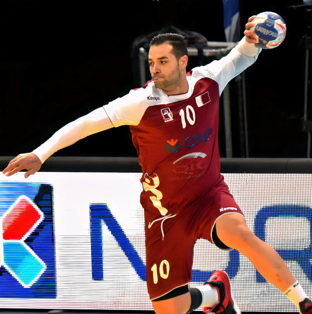 Ein Mann in einer rot-weißen Uniform, der Handball spielt und einen Ball hält, mit entschlossener Miene und ausgestreckten Armen vor einem "QNP Handball World Cup 2019"-Schild.