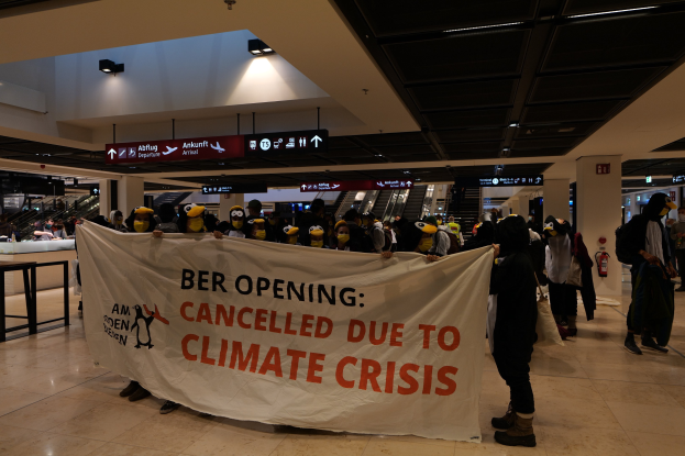 Eine Gruppe von Menschen in Kostümen hält ein Banner mit der Aufschrift 'Beröffnung abgesagt aufgrund der Klimakrise' in einem Flughafen mit Tischen, Stühlen, Rolltreppen und Informationsanzeigen im Hintergrund.