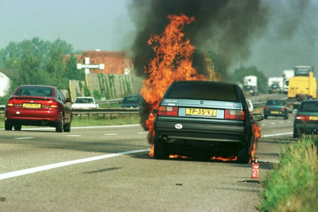 Ein Auto, das in Flammen steht, mit anderen Fahrzeugen in der Nähe, Bäumen, Gebäuden und einem klaren blauen Himmel im Hintergrund und Gras mit einem Feuerlöscher auf der rechten Seite.