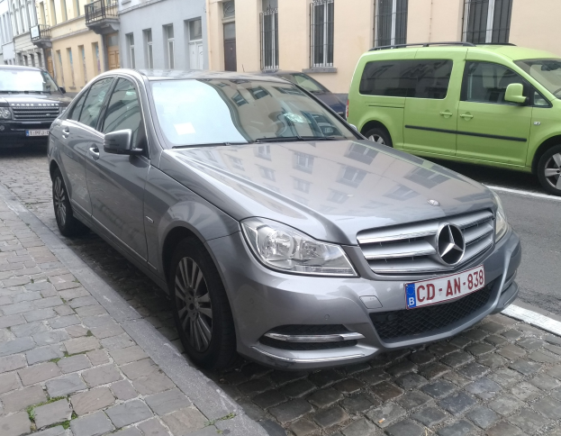 Ein Mercedes-Benz C 220 CDI BlueEfficiency Avantgarde fährt auf einer Straße, die von Gebäuden mit Fenstern, Geländern und Namensschildern gesäumt ist, unter einem sichtbaren Himmel.