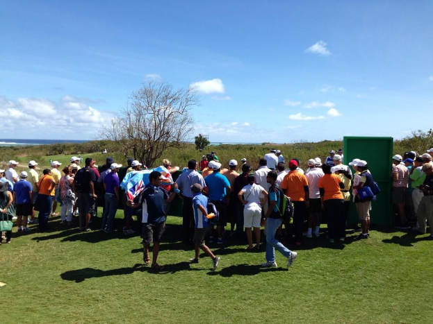 Gruppe von Menschen auf einem grünen Feld, die ein Golfturnier beobachten, mit Bäumen, Pflanzen, einer bewölkten Himmel und einem Gewässer im Hintergrund.