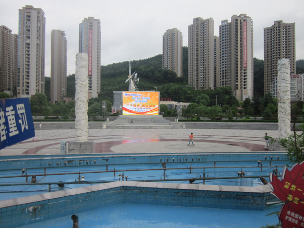 Großer Swimmingpool umgeben von hohen Gebäuden mit Menschen drumherum, eine Fahne links, Bäume im Hintergrund und einen klaren blauen Himmel.