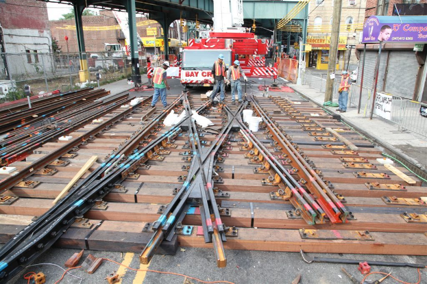 Arbeiter in Jacken und Mützen bauen Schienen, mit einem Fahrzeug mit Materialien im Hintergrund und einer Hütte mit Pfählen daneben und einem Geschäft auf der anderen Seite.