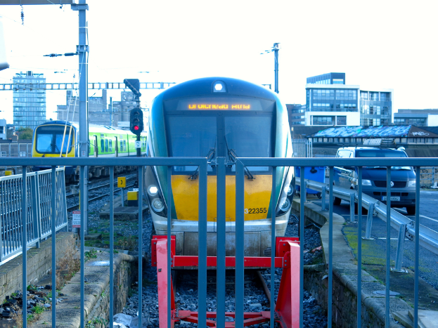 Zwei Züge auf parallelen Gleisen mit einem Zaun im Vordergrund, ein Auto auf der Straße rechts, eine Signalleuchte auf einem Pfahl, Gebäude im Hintergrund und ein Himmel darüber.