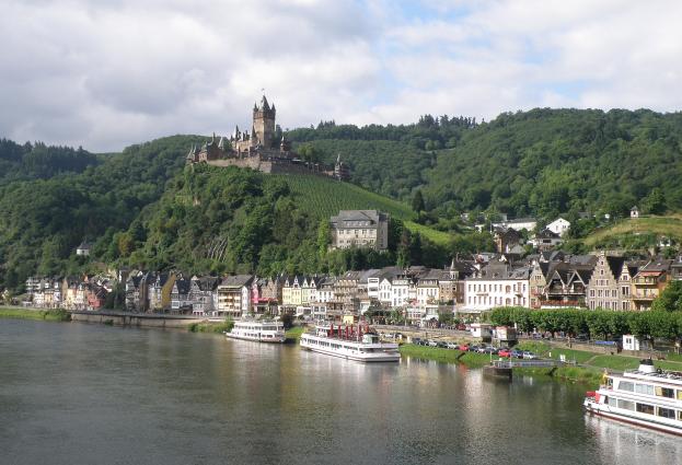Eine malerische Aussicht auf den Rhein in Deutschland, mit einer Burg auf einem Hügel, Booten auf dem Wasser, Fahrzeugen auf der Straße und einer bewölkten Himmel.