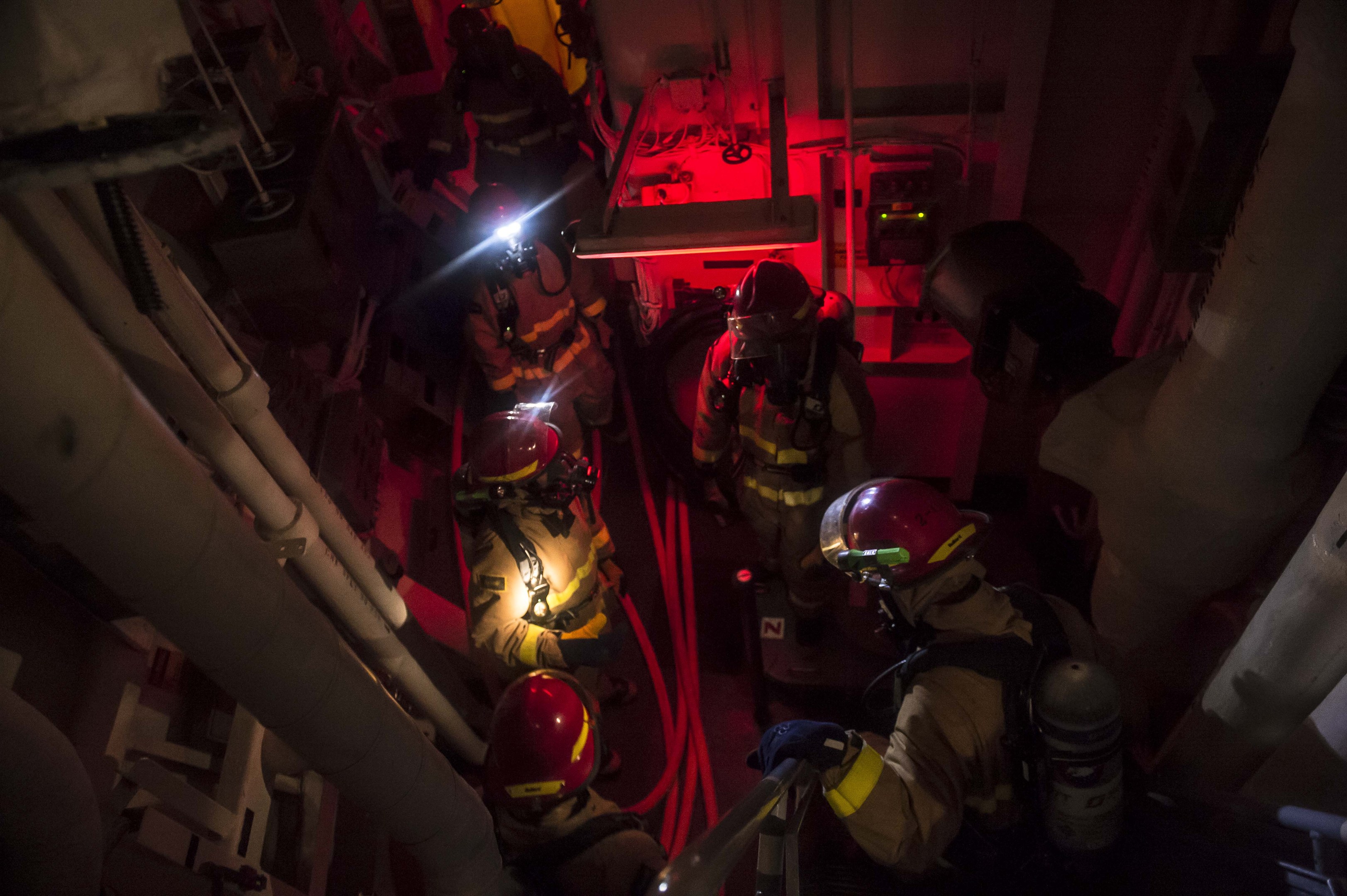 Feuerwehrleute in Helmen und Sauerstoffflaschen stehen in einem schwach beleuchteten Raum mit Rohren auf beiden Seiten und bereiten sich auf einen Rettungseinsatz in der Nähe von Maschinen und anderer Ausrüstung vor.