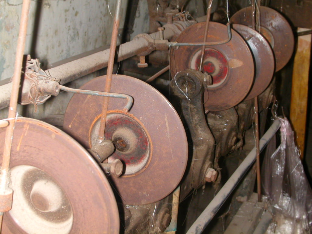 Ein Nahaufnahme eines Metall-Schleifmaschinen mit einem großen zentralen Rad und mehreren kleineren umgebenden Rädern, mit einem Seitengriff, vor einem hellgrauen, schmutzigen Wand.