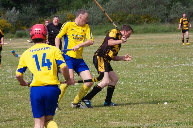 Menschen spielen Hockey auf Wiese, mit anderen Menschen und Grünfläche im Hintergrund.