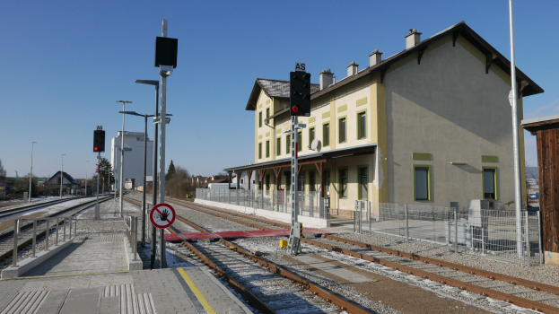 Ein Bahnhof mit einem Zug auf den Schienen, umgeben von Gebäuden, Straßenlaternen, Verkehrspfosten, Verkehrsampeln, Schildern, Zäunen, Bäumen und einem klaren blauen Himmel.