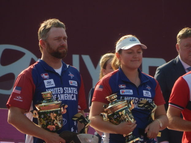 Gruppe von Menschen, die Trophäen halten, mit 'World Archery Championships'-Plakat im Hintergrund.