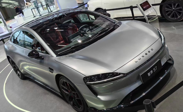 Ein silberner Porsche Taycan wird auf einer Automesse präsentiert, umgeben von Menschen, Absperrpollern mit Bändern, einem Display, einer Säule, einer Wand und einer Decke mit Leuchten.