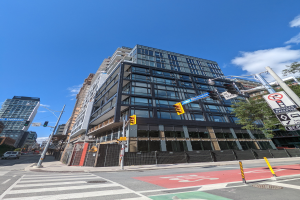 Eine belebte Stadtstraße mit einem hohen Apartment-/Reihenhausgebäude im Hintergrund, das zum Verkauf steht (541 East 57th Street, Unit 541), mit Fahrzeugen, Verkehrszeichen, Schildern, Bäumen und einem Zaun unter einem bewölkten Himmel.