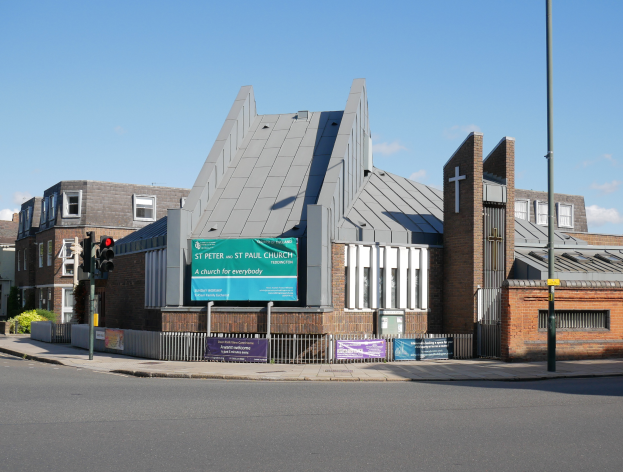 St. Peter und St. Paul Kirche in London, England, mit Fenstern, einem Metallzaun, Bannern mit Text, einem Schild, einem Straßenpfahl, einem Verkehrssignal, einigen Pflanzen und einem bewölkten Himmel.