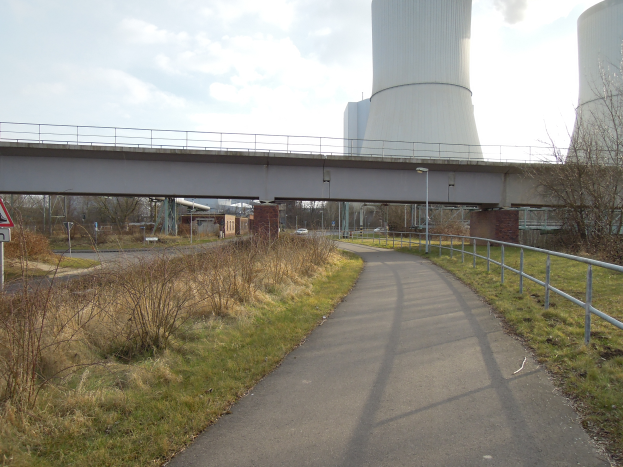 Eine von Gras und Pflanzen gesäumte Straße führt zu einer Brücke mit Geländern, Laternenmästen und einem Schild, im Hintergrund ein Atomkraftwerk mit Rauch aus dem Schornstein unter einem bewölktem Himmel.