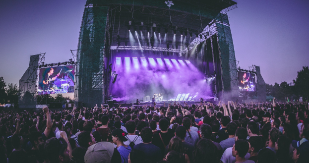 Eine große Menschenmenge auf einem Musikfestival schaut auf einer beleuchteten Bühne mit Instrumenten und Bildschirmen, die vor einem Gebäude, Bäumen und einem klaren Himmel stehen.