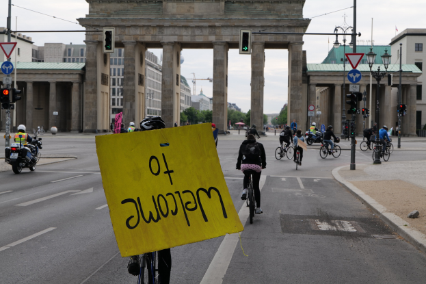Eine Gruppe von Menschen auf Fahrrädern fährt eine Straße in Berlin, Deutschland, vor dem Brandenburger Tor entlang, trägt Helme und hält ein gelbes Schild hoch, mit Laternenpfählen, Verkehrsampeln, Gebäuden, Bäumen und einem klaren blauen Himmel im Hintergrund.