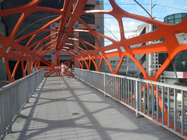 Oranger Metallfußgängerbrücke mit Menschen darauf, die eine Stadtstraße überqueren, mit Gebäuden, Strommasten und Drähten im Hintergrund unter einem klaren blauen Himmel.