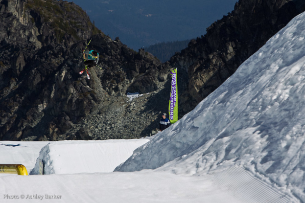 Ein Mann in Skier springt in die Luft mit schneebedeckten Bergen und einer Flagge mit Text im Hintergrund und einem anderen Mann vor der Flagge mit Text in der unteren linken Ecke.