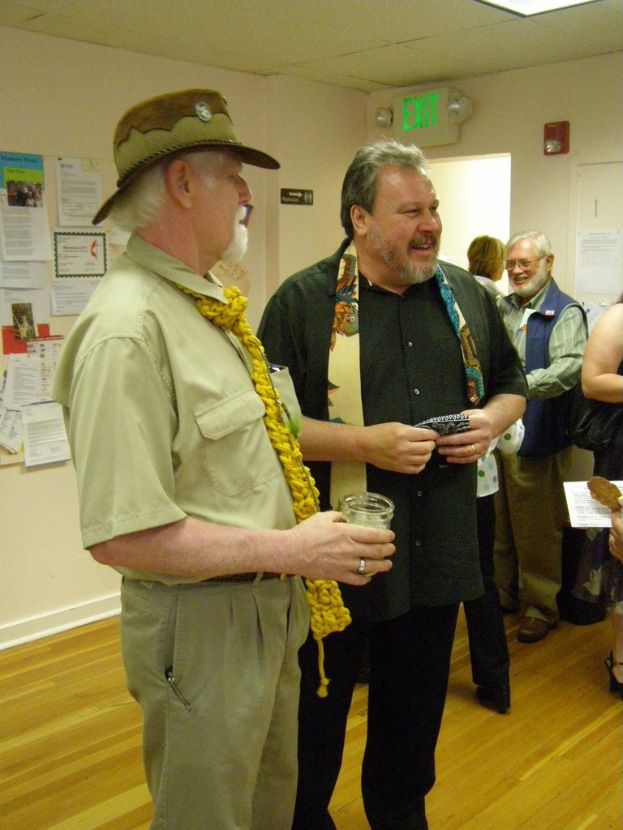 Eine Gruppe älterer Männer, die auf einem Holzfußboden in einem Raum mit Papieren an den Wänden, einer Warntafel an der rechten Wand, einer Tür darunter und Deckenleuchten steht.