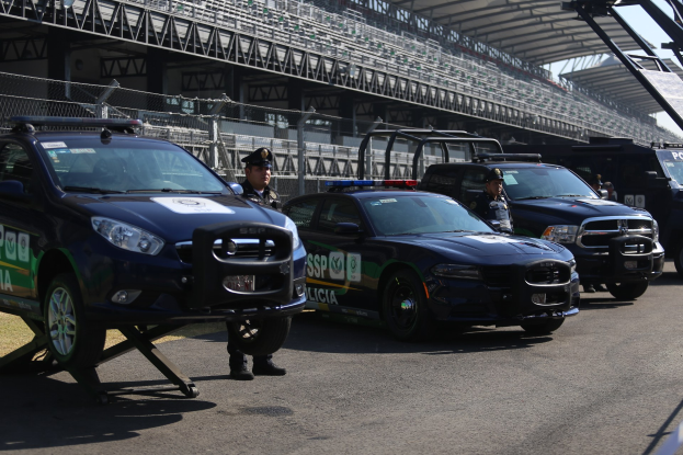Polizeiautos vor einer Rennstrecke geparkt mit uniformierten Personen, ein Stadion mit Geländern und Maschendrahtzaun im Hintergrund.
