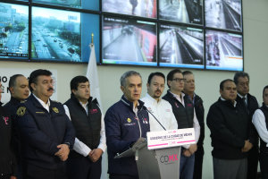 Eine Gruppe von Männern steht vor einem Rednerpult mit einem Mikrofon und einer Tafel mit der Aufschrift "Mexikanisches Zoll- und Grenzschutz". Im Hintergrund sind Fernseher und eine Flagge zu sehen.