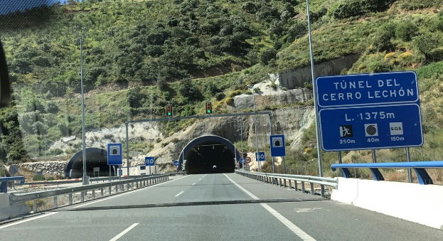 Auto fährt durch einen Tunnel auf einer Straße mit Geländern, Schildern, Pfählen, Ampeln, Bäumen, Felsen und einem klaren blauen Himmel.