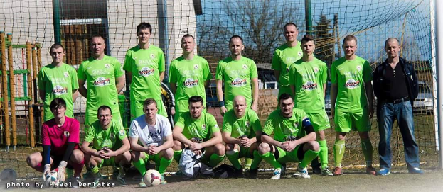 Eine Gruppe von Männern in grünen T-Shirts steht und hockt auf einem Fußballfeld, mit einem Ball vor ihnen und einem Torpfosten rechts daneben, und einem Hintergrund aus Bäumen, Gebäuden, Pfählen, Fahrzeugen und dem Himmel.