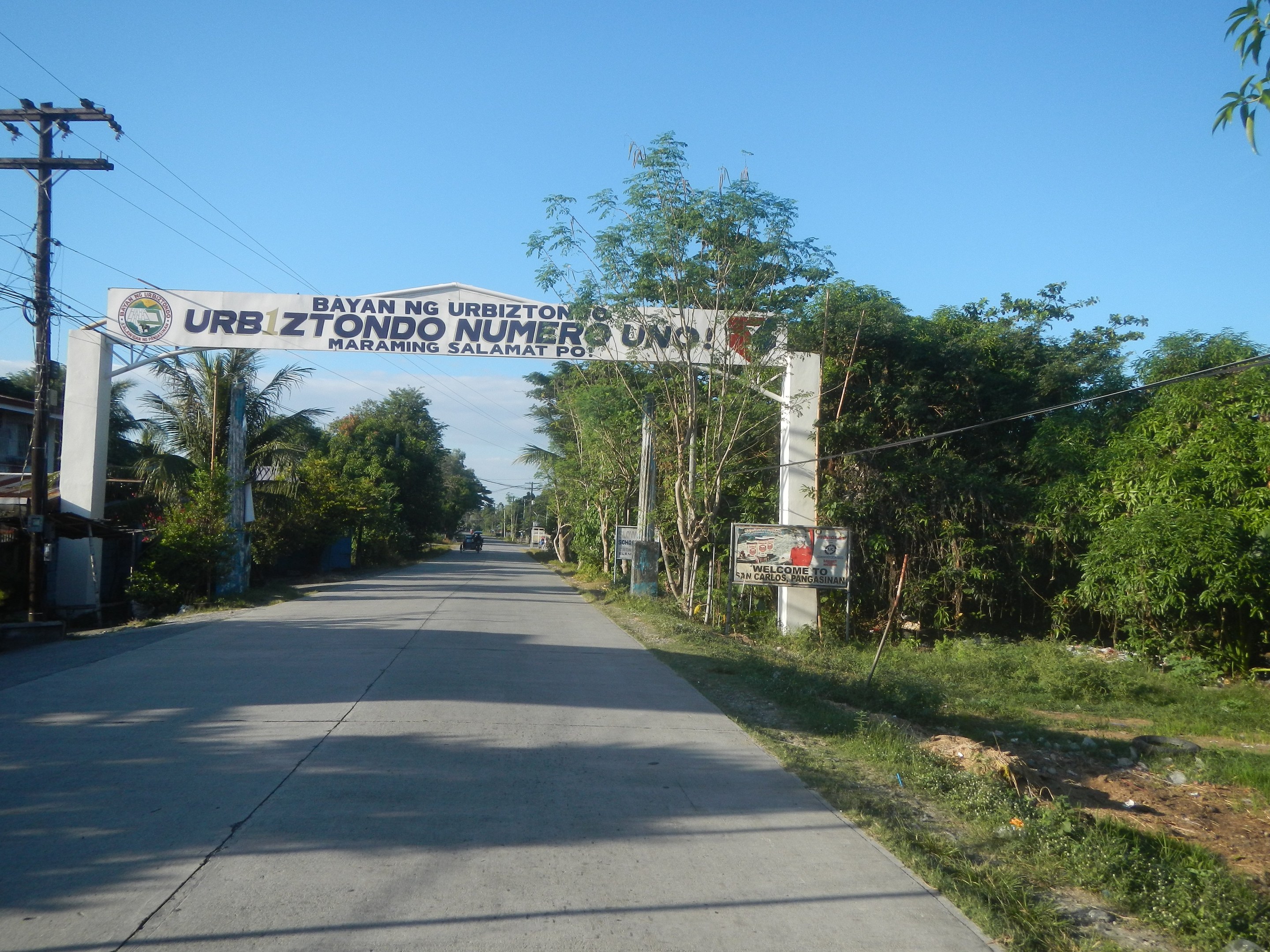Straßenbogen mit "Zu verkaufen Grundstück / Land / Farm Cavite > Imus 1"-Text, flankiert von Bäumen, Strommasten mit Drähten, Grasboden und Himmel im Hintergrund.