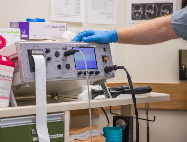 Eine Person in blauen Handschuhen arbeitet an einem medizinischen Gerät auf einem Tisch, der mit verschiedenen Gegenständen überhäufte ist, mit Papieren an der Wand im Hintergrund, was auf eine Krankenhausumgebung hindeutet.