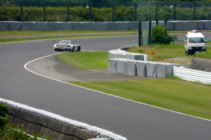 Ein Rennwagen fährt eine Rennstrecke entlang, im Hintergrund ist ein Polizeiwagen zu sehen, umgeben von Gras, Pflanzen, Bäumen und Absperrungen, mit einem Fahrzeug auf der rechten Seite und Text am unteren Rand.