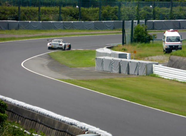 Ein Rennwagen fährt eine Rennstrecke entlang, im Hintergrund ist ein Polizeiwagen zu sehen, umgeben von Gras, Pflanzen, Bäumen und Absperrungen, mit einem Fahrzeug auf der rechten Seite und Text am unteren Rand.