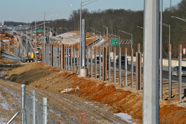 Baustelle mit Fahrzeugen, Masten, Beleuchtung, Beschilderung, einem Zaun, schneebedecktem Gras, Bäumen und einem Himmel im Hintergrund.