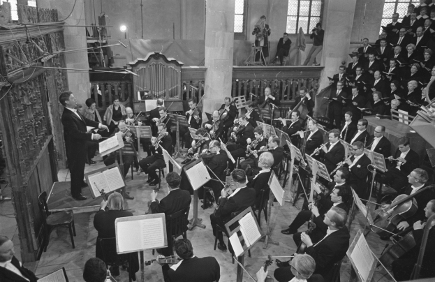 Schwarzes und weißes Bild einer Gruppe von Menschen, die musikalische Instrumente in einer Kirche spielen, die auf Stühlen sitzen und auf dem Boden stehen mit Notenpulten vor ihnen.