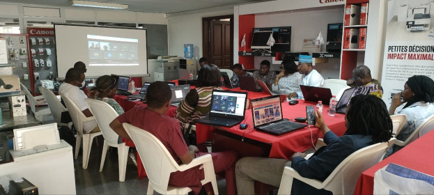 Gruppe von Menschen, die um einen Tisch mit Laptops und anderen Gegenständen während eines digitalen Marketing-Workshops sitzen, mit einem Banner, einem Bildschirm, einer Tür und Deckenleuchten im Hintergrund.