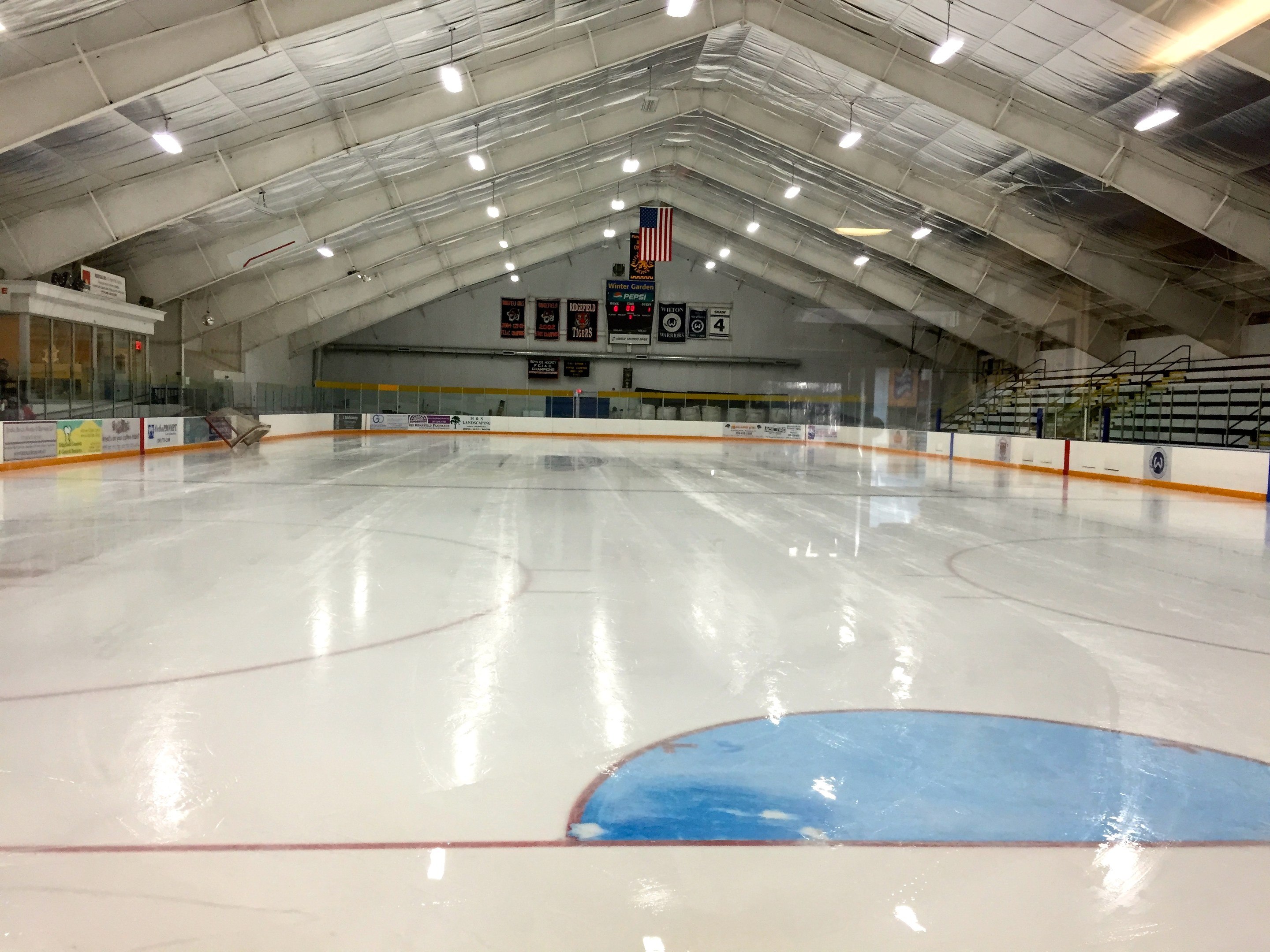 Große Indoor-Eisbahn mit Deckenbeleuchtung, Werbetafeln, einer Wand im Hintergrund, Treppen mit Geländern und einigen Menschen, was auf eine Sportanlage hinweist, die likely für Hockey verwendet wird.