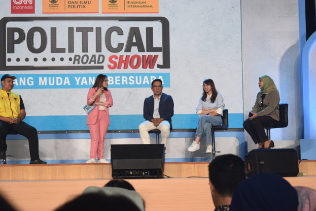 Eine Gruppe von Menschen auf einer Bühne sitzend, mit einer Frau in der Mitte, die ein Mikrofon hält, und einem Publikum im Vordergrund, mit einem Banner mit der Aufschrift "Politischer Roadshow 2019" im Hintergrund.