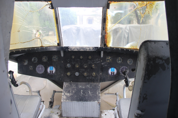 Cockpit-Innenseite eines Flugzeugs mit zerbrochener Frontscheibe, zwei Sitzen und mehreren Kontrollinstrumenten, die Bäume und ein Gebäude äußer zeigen.