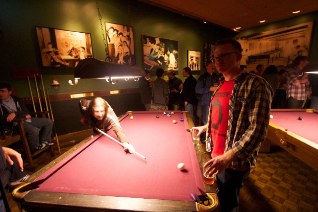 Zwei Personen spielen Snooker an einem von zwei Tischen in einem Club, während mehrere andere im Hintergrund stehen und reden, vor einer cremefarbenen Wand mit Fotopostern.
