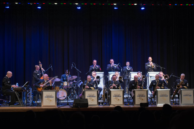 Eine Gruppe von Musikern auf der Bühne, die Instrumente spielen, mit Mikrofonen und Ständern, Schildern mit der Aufschrift "The New Orleans Jazz Band", ein Vorhang im Hintergrund und ein Publikum darunter unter Bühnenlichtern.