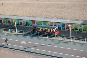 Ein kleiner Zug mit Passagieren fährt auf Schienen neben einem Strand, mit einer Person zu Fuß auf dem linken Fußweg, einer Wand mit Plakaten im Hintergrund und sichtbarem Boden am oberen Bildrand.