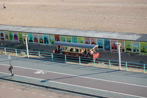 Ein kleiner Zug mit Passagieren fährt auf Schienen neben einem Strand, mit einer Person zu Fuß auf dem linken Fußweg, einer Wand mit Plakaten im Hintergrund und sichtbarem Boden am oberen Bildrand.
