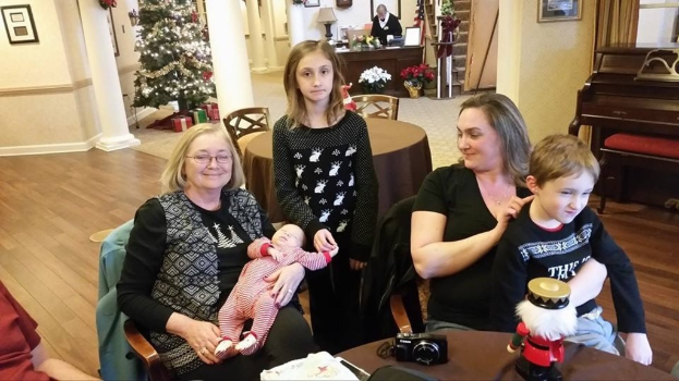 Ein Raum mit einer Wand, die ein Foto Display, einen Weihnachtsbaum, eine Lampe und Pflanzen mit Blumen zeigt. Es gibt Treppen, einige Menschen, die auf Stühlen und Tischen sitzen, mit einem Spielzeug und einer Kamera auf einem der Tische.