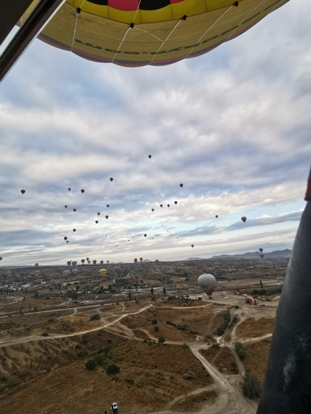 Eine Gruppe von Heißluftballons, die über eine Wüstenlandschaft fliegen, mit Fallschirmen am Himmel und einem Fahrzeug auf dem Boden darunter, unter einem bewölkten Himmel.