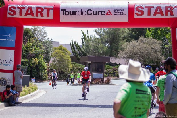 Eine belebte Straßenszene mit Menschen auf Fahrrädern, einige sitzen auf einem Gehweg, andere stehen auf der Straße, mit einer Hütte und umliegenden Pflanzen und Bäumen im Hintergrund.