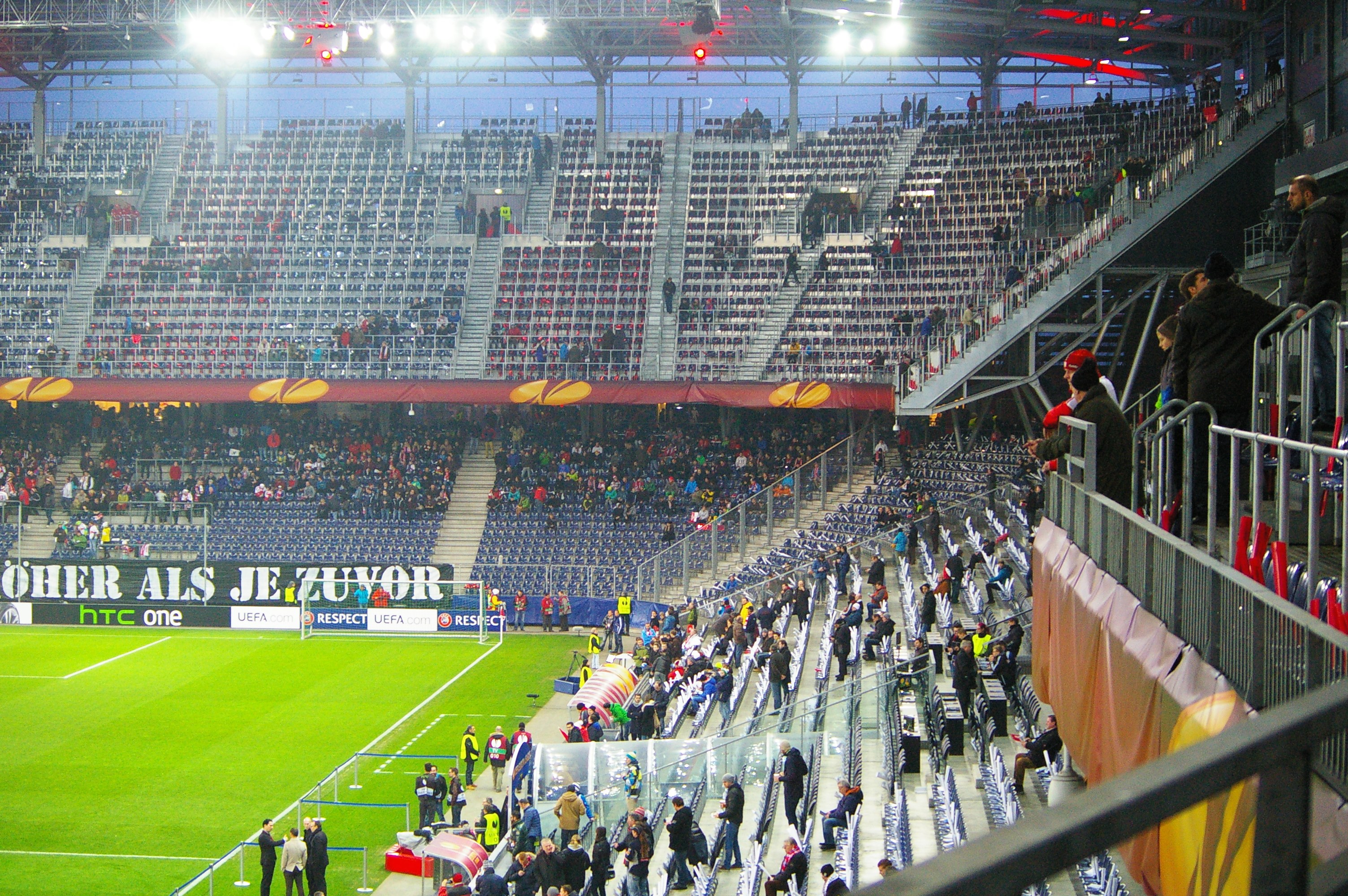 Großes Stadion voller Zuschauer bei einem Fußballspiel, mit sitzenden und stehenden Personen, einem Geländer, einer rechten Banner und einem leeren beleuchteten Bereich im Hintergrund.