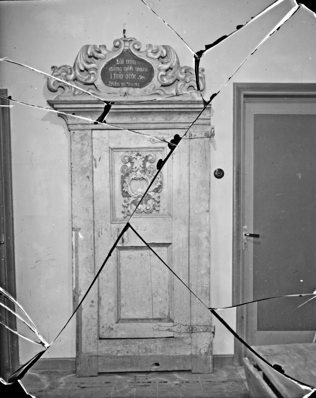 Ein Schwarz-Weiß-Foto einer kaputten Tür mit verstreuten Glasscherben in einem Raum mit einer Wand im Hintergrund.