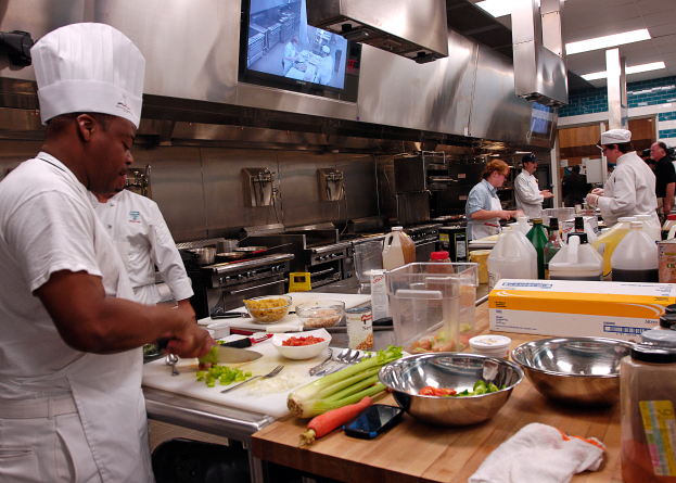 Eine Gruppe von Köchen in einer Küche, die Essen auf einem Schneidbrett zubereiten, mit einem Koch im Vordergrund in einer weißen Schürze und einer Mütze, der ein Messer hält und von verschiedenen Utensilien, Schüsseln, Gemüse, einem Handy, Flaschen und anderen Gegenständen umgeben ist, mit einem Fernseher und Deckenleuchten im Hintergrund.