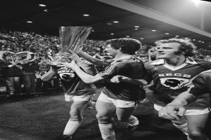 Eine Gruppe von Männern, die einen Pokal auf einem Fußballfeld halten und ihren FA-Cup-Sieg feiern, umgeben von einer Menge hinter einem Zaun, unter Stadionlichtern, in Schwarz-Weiß.